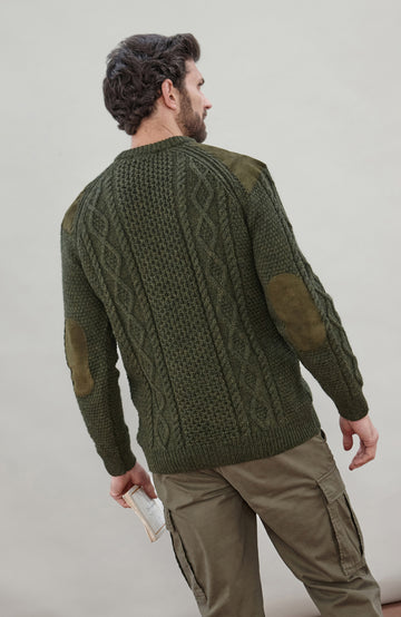 A man shown from the back wearing an olive green cable-knit sweater with brown elbow patches, paired with khaki cargo pants, against a neutral light gray background.