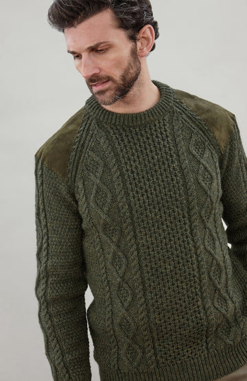 A man wearing a dark olive green cable-knit made in England sweater by Paul James Knitwear with contrasting dark olive shoulder panels, photographed against a plain light gray studio background.
