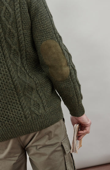 A man wearing a dark olive green cable-knit made in England sweater by Paul James Knitwear