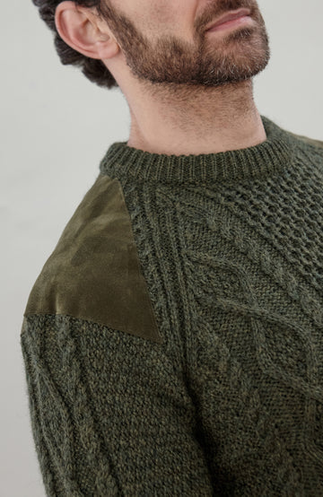 Close-up of a man wearing an olive green cable-knit sweater with a contrasting solid olive green suede shoulder panel, shown from the chest up against a light gray background.