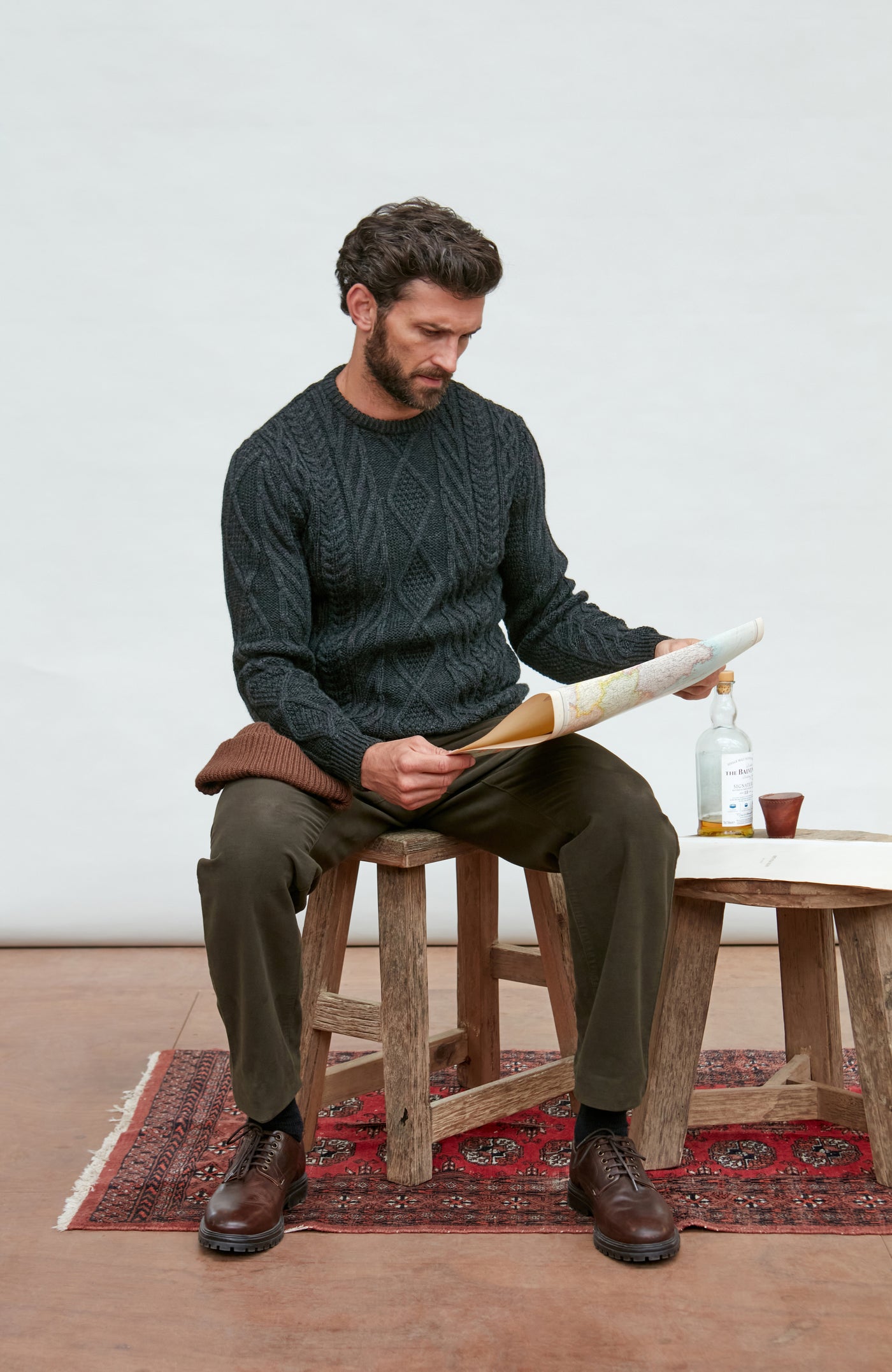 Man sitting on a stool holding a shoe, wearing a dark green sweater and brown pants, with a white wall and wooden table in the background.