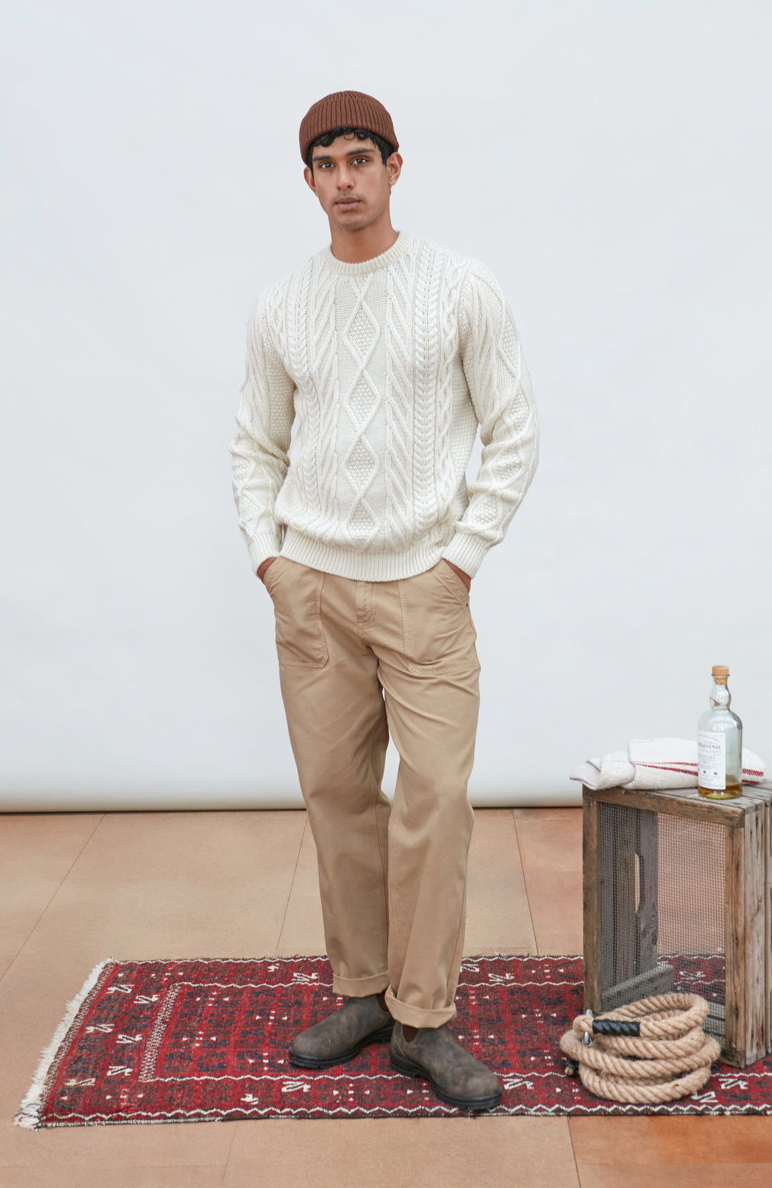 A man wearing a cream merino wool cable-knit crewneck sweater made in england by Paul James Knitwear, tan pants, and a brown beanie hat, standing against a light gray studio background.