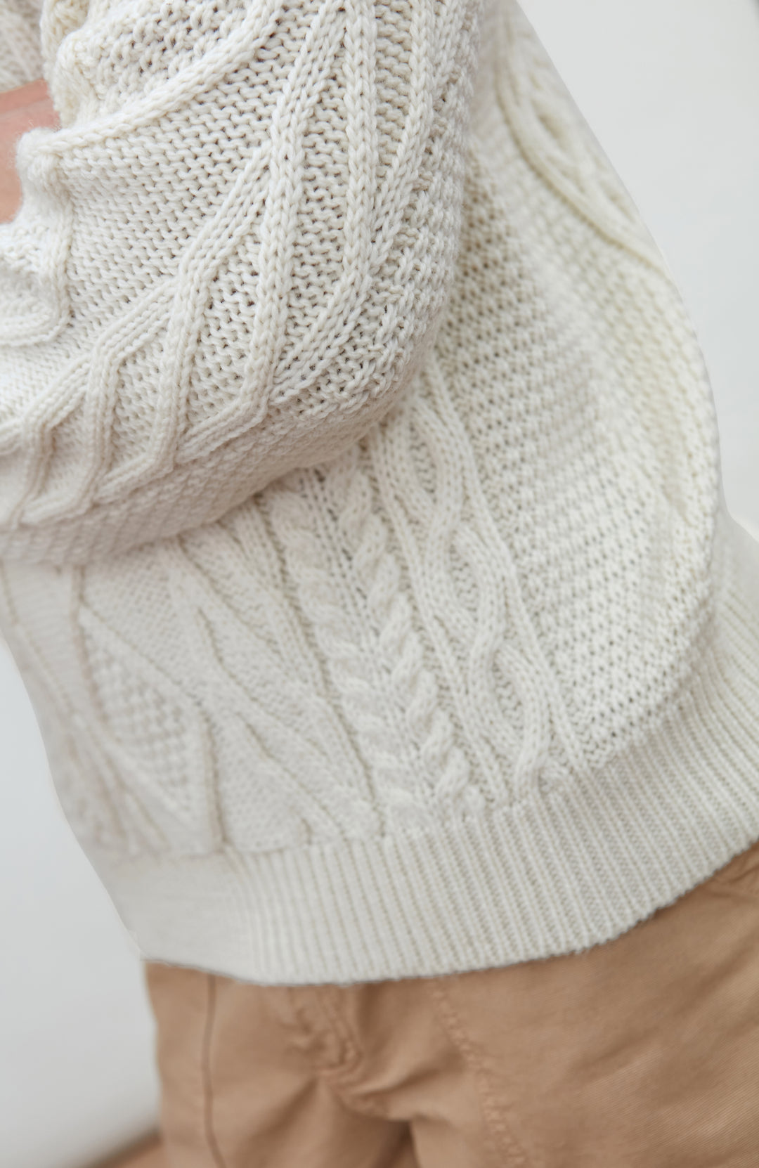 A man wearing a cream merino wool cable-knit crewneck sweater made in england by Paul James Knitwear, tan pants, and a brown beanie hat, standing against a light gray studio background.