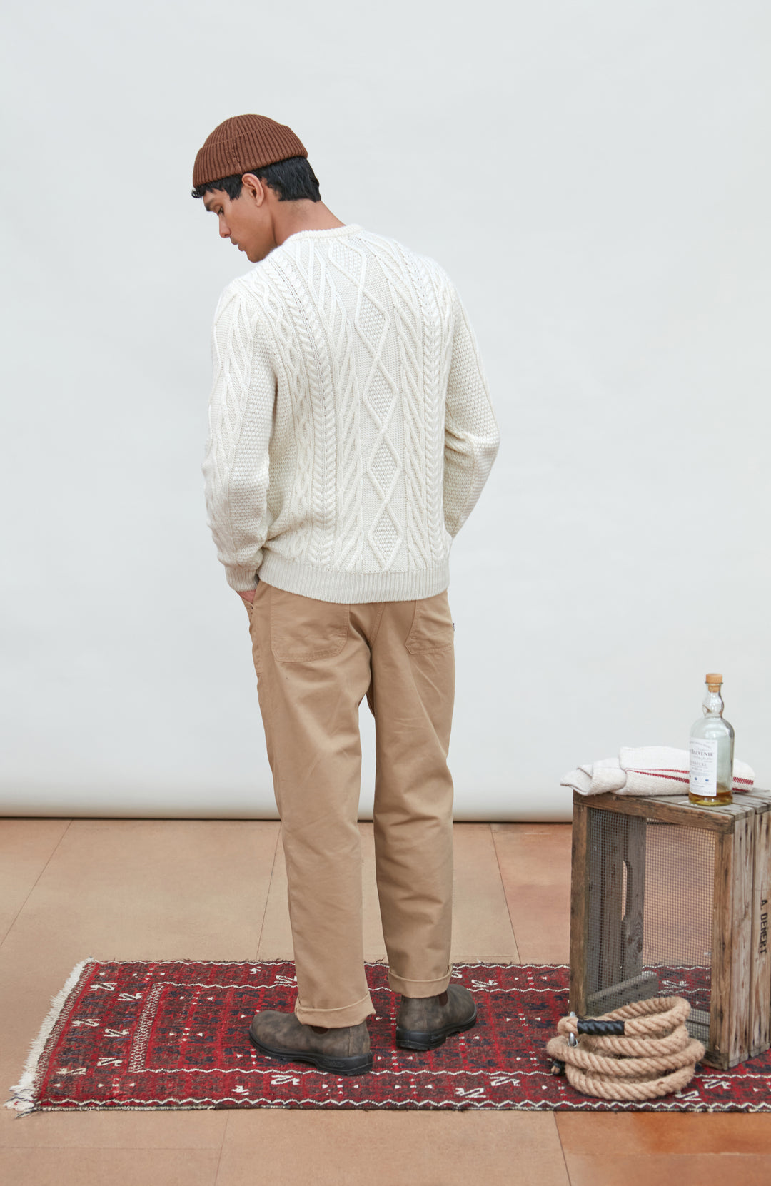 A man wearing a cream merino wool cable-knit crewneck sweater made in england by Paul James Knitwear, tan pants, and a brown beanie hat, standing against a light gray studio background.