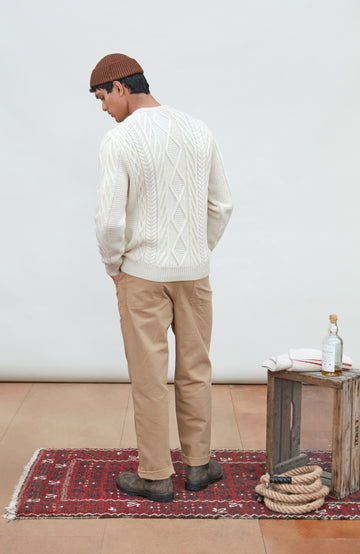 A man wearing a cream merino wool cable-knit crewneck sweater made in england by Paul James Knitwear, tan pants, and a brown beanie hat, standing against a light gray studio background.