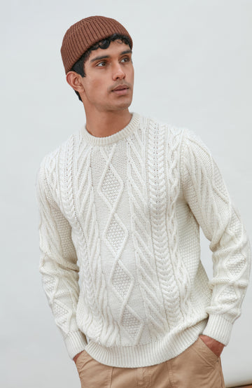 A man wearing a cream merino wool cable-knit crewneck sweater made in england by Paul James Knitwear, tan pants, and a brown beanie hat, standing against a light gray studio background.