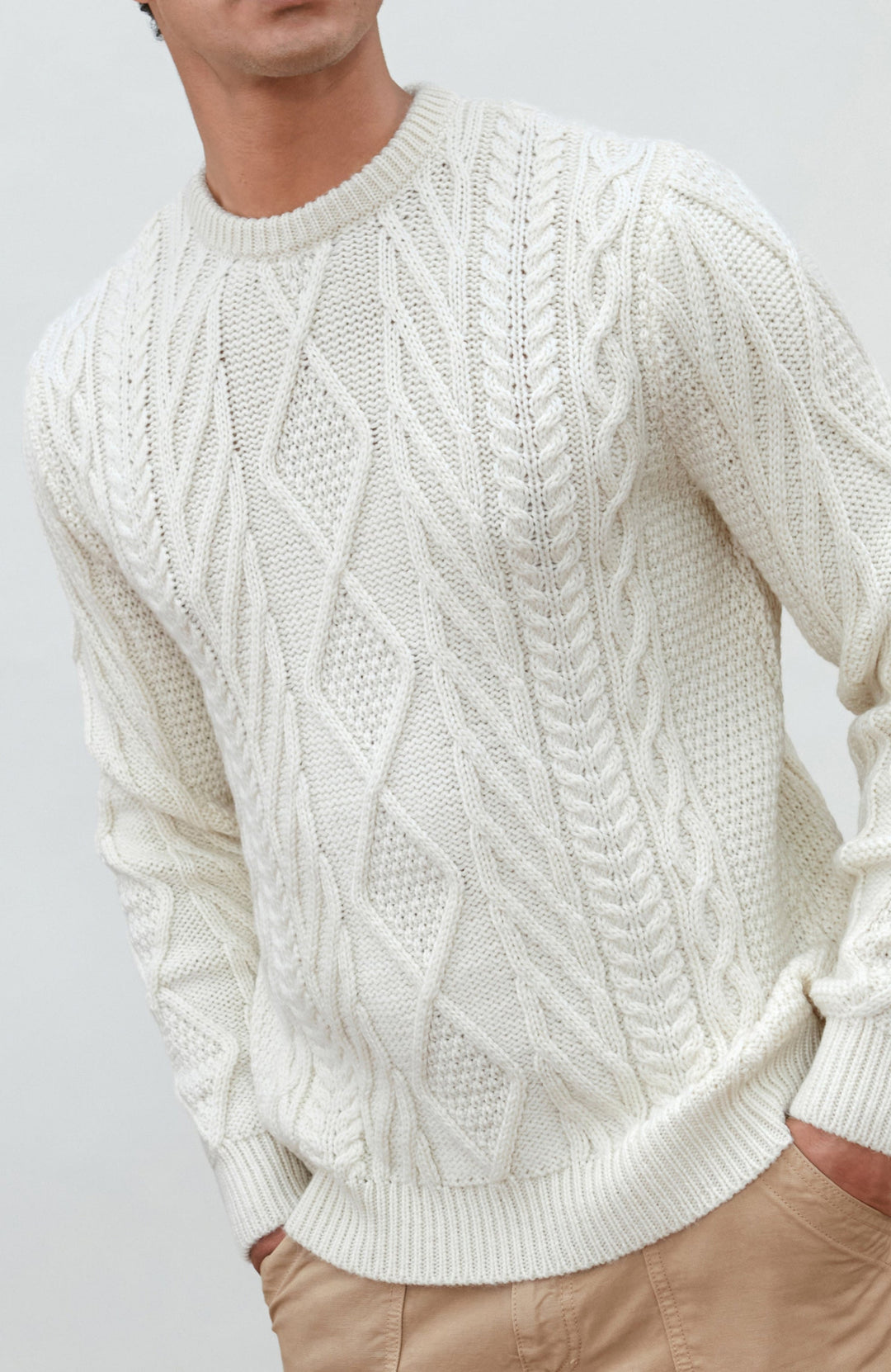 A man wearing a cream merino wool cable-knit crewneck sweater made in england by Paul James Knitwear, tan pants, and a brown beanie hat, standing against a light gray studio background.