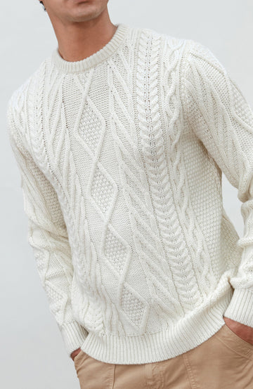 A man wearing a cream merino wool cable-knit crewneck sweater made in england by Paul James Knitwear, tan pants, and a brown beanie hat, standing against a light gray studio background.