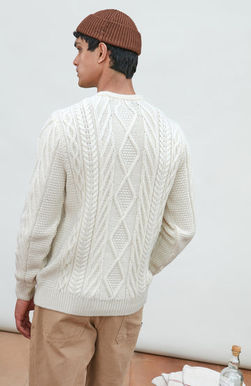 A man wearing a cream merino wool cable-knit crewneck sweater made in england by Paul James Knitwear, tan pants, and a brown beanie hat, standing against a light gray studio background.