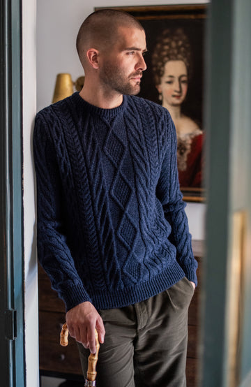 Man wearing a blue cable wool knit sweater standing in a room with a painting in the background by Paul James