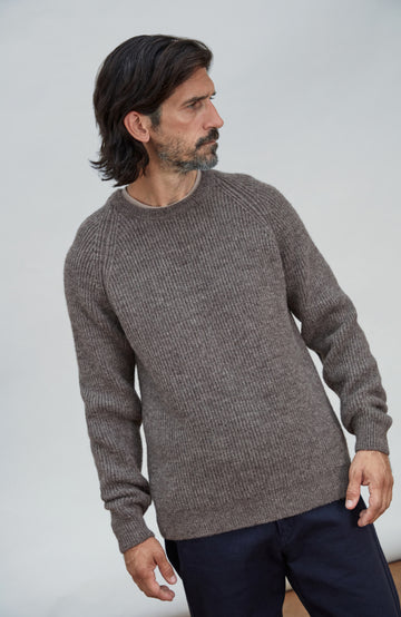 Made in England men's wool rib fisherman jumper. A man wearing a brown ribbed knit crewneck sweater and dark navy trousers, standing against a light gray studio background.