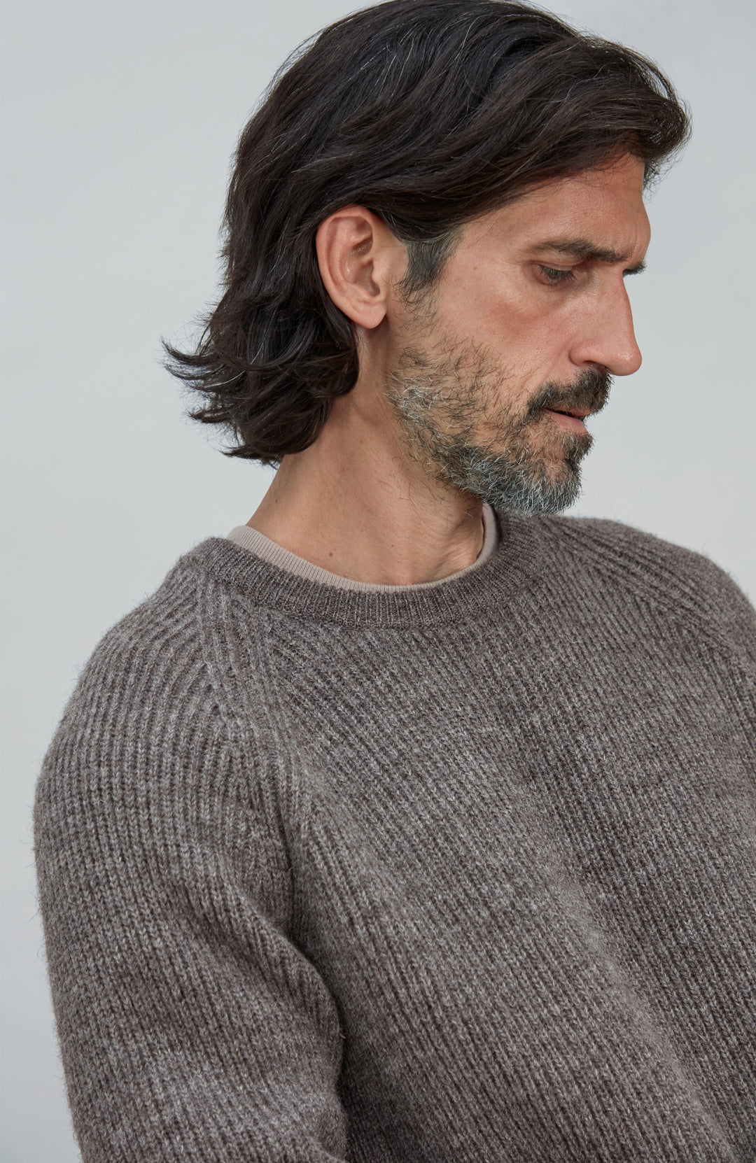 A man with dark hair and a beard wearing a textured, ribbed knit crewneck sweater in a warm taupe-brown color, shown in a close-up profile shot against a light gray background. Made in England men's wool rib fisherman jumper.