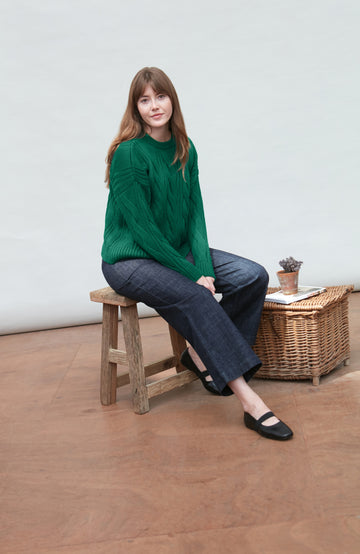 Woman sitting on a wooden stool wearing a green chunky wool paul james sweater and dark pants in a minimalistic room.