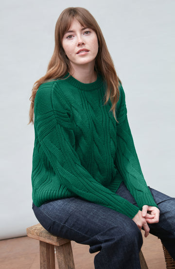 Woman wearing a green chunky wool paul james sweater sitting on a wooden stool against a plain background