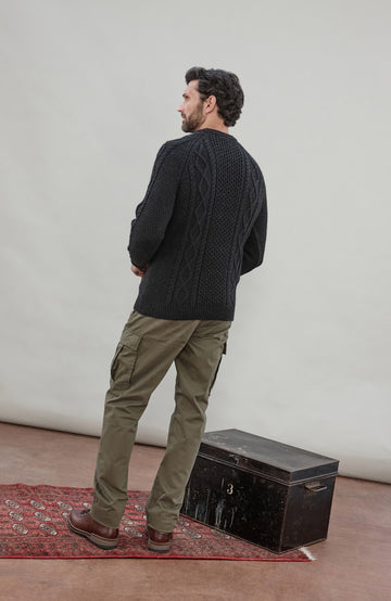 A man stands with his back to the camera wearing a dark charcoal cable-knit sweater, olive green cargo pants, and brown boots, standing next to a vintage black trunk on a red patterned rug against a light gray studio backdrop.