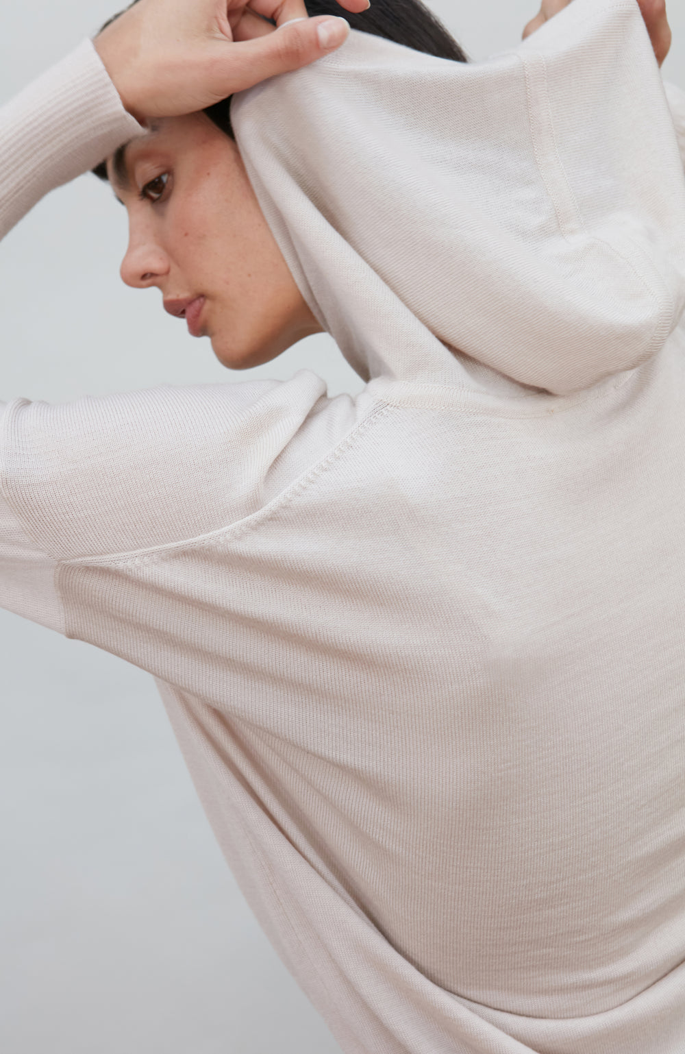 A woman wearing a soft, light beige merino wool knit hooded sweater by paul james knitwear against a plain light gray background.