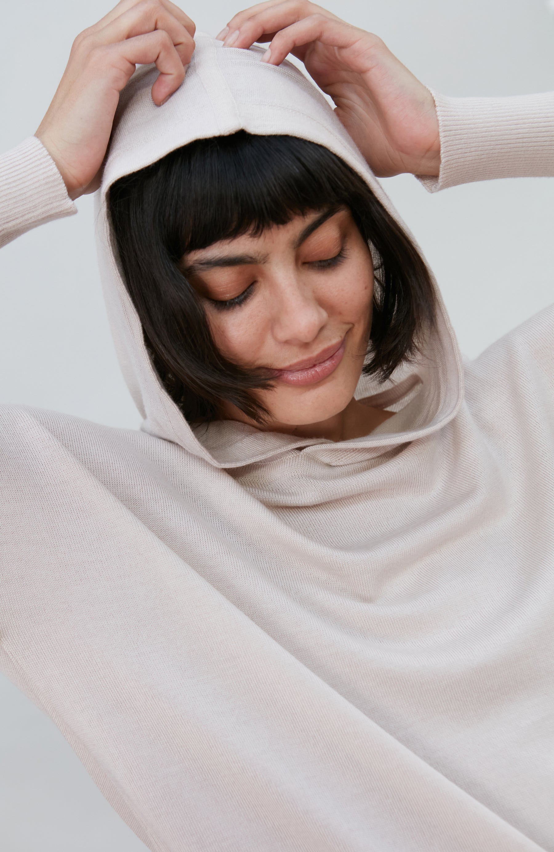 A woman wearing a soft, light beige merino wool knit hooded sweater by paul james knitwear against a plain light gray background.