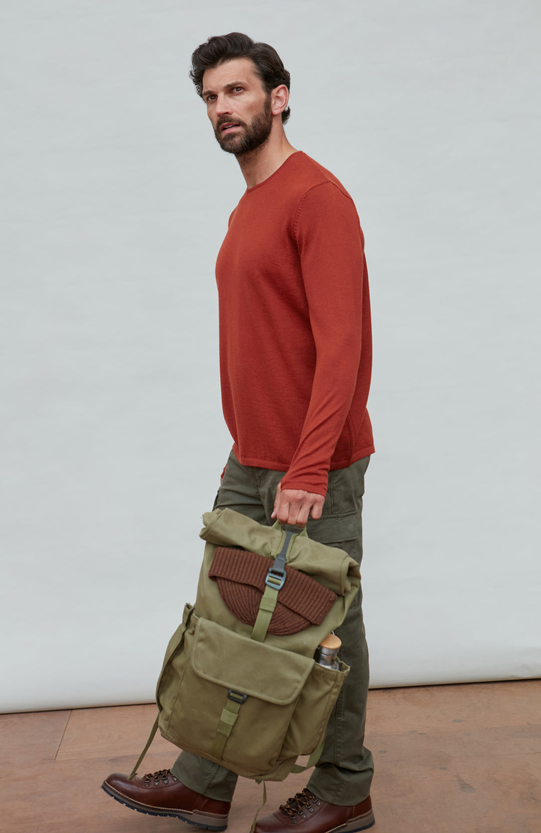 Man in wool jumper holding a green backpack against a plain background by Paul James