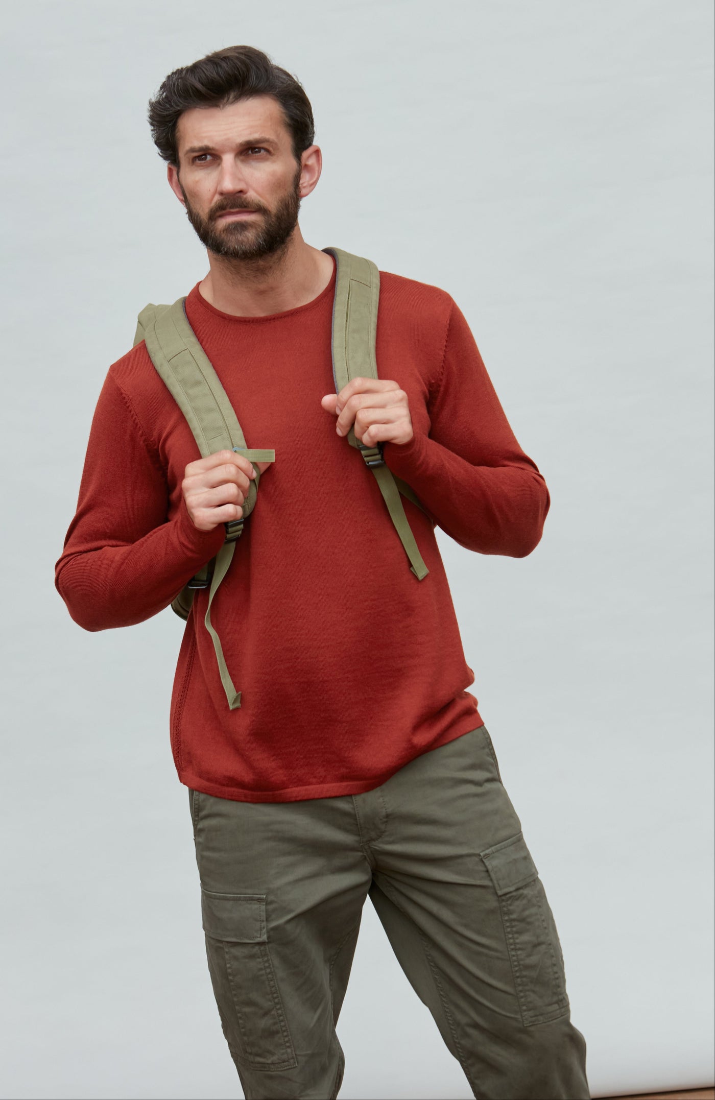 Man wearing a red merino wool sweater and green backpack against a plain background