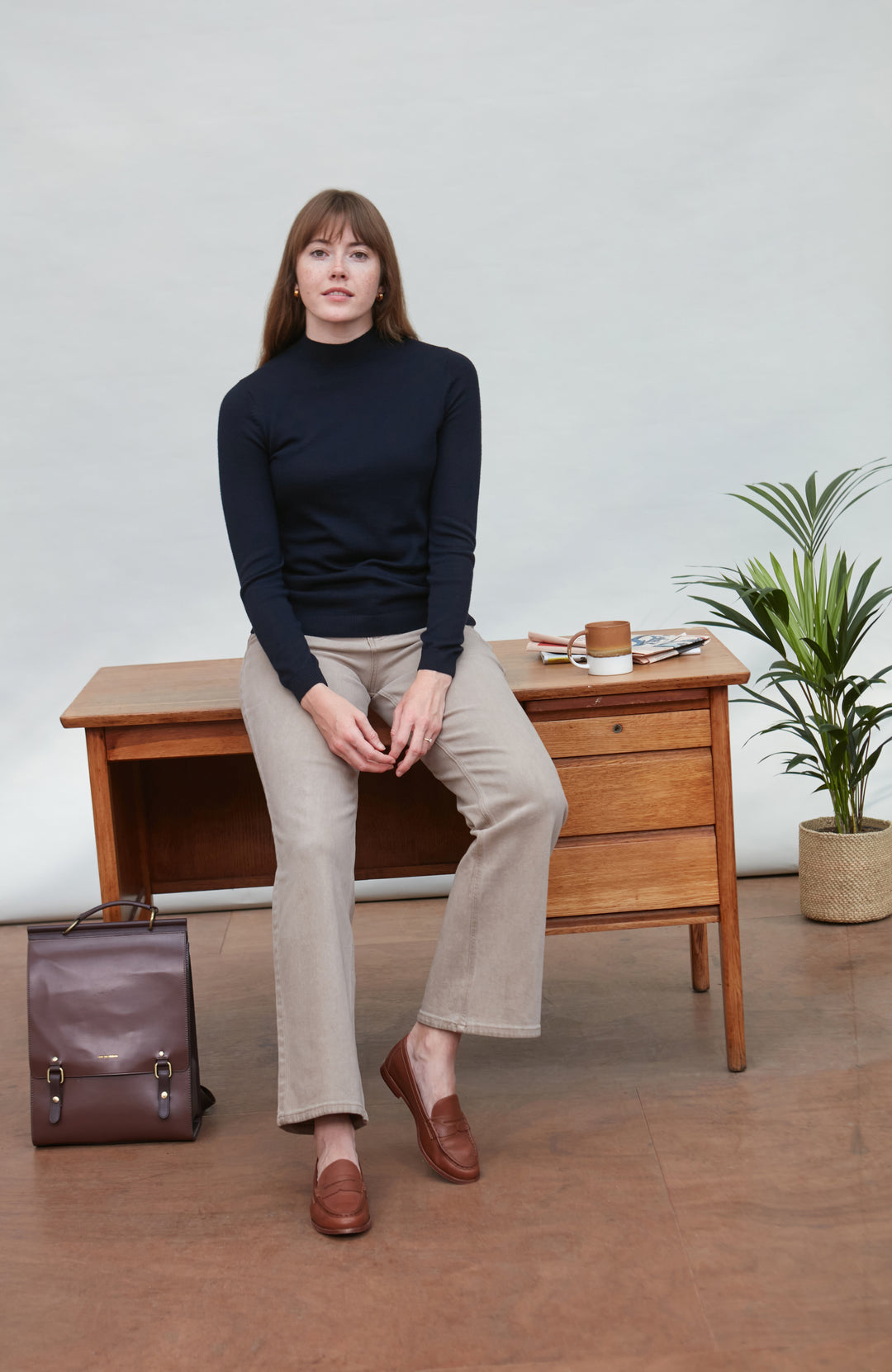 Woman sitting on a wooden desk wearing a navy turtleneck, beige pants, and brown shoes in a minimalistic room from paul james