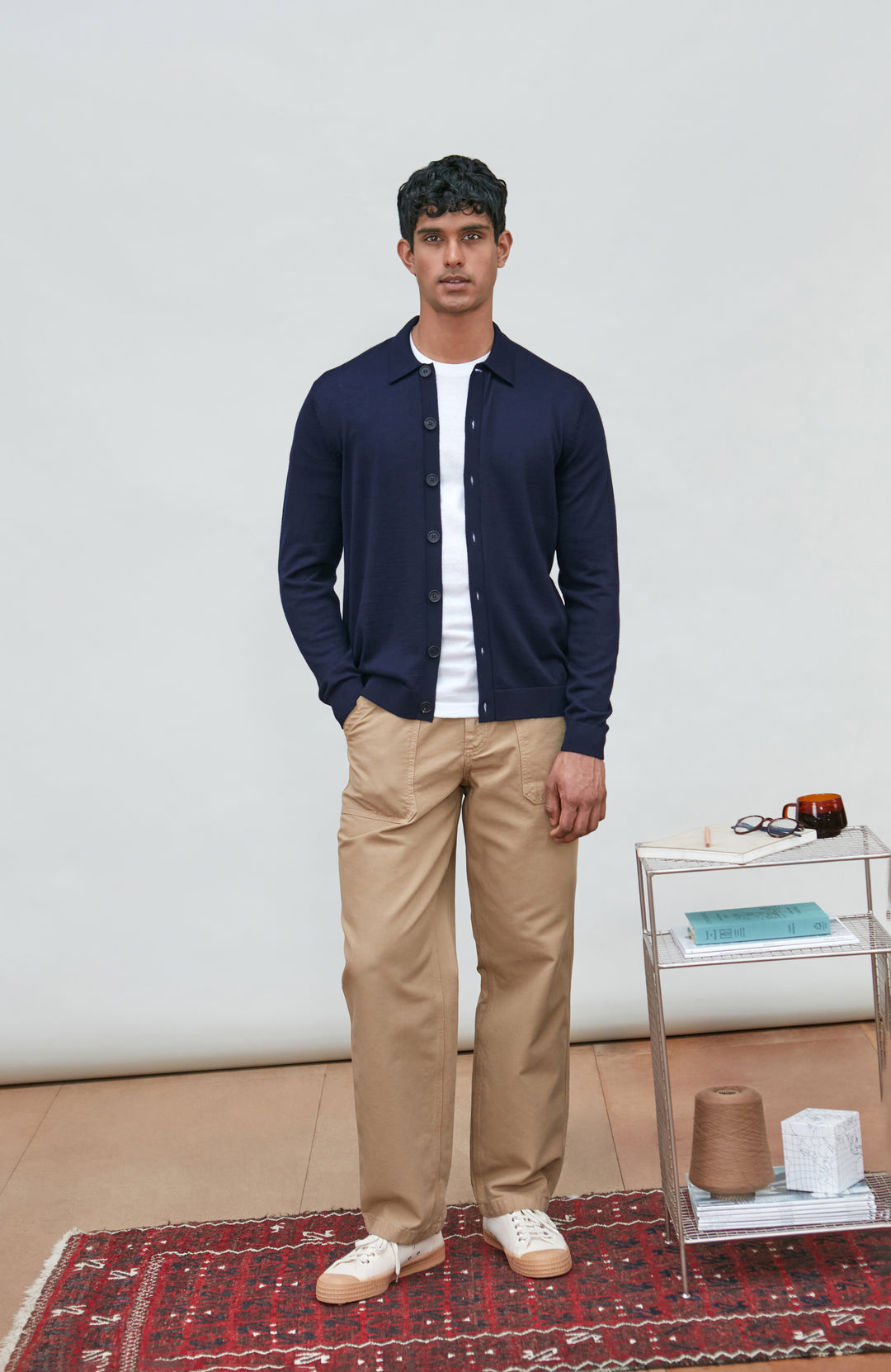 A man wearing a navy blue fine merino wool polo button-up knit cardigan by paul james knitwear over a white undershirt and khaki pants, standing against a plain light gray studio background.