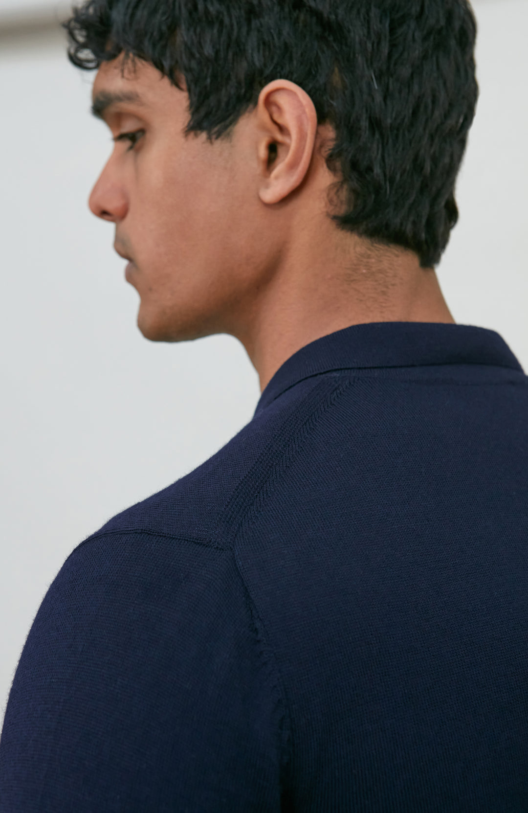 A man wearing a navy blue fine merino wool polo button-up knit cardigan by paul james knitwear over a white undershirt and khaki pants, standing against a plain light gray studio background.