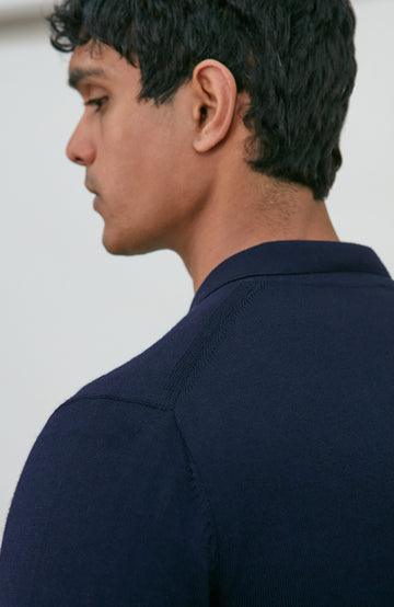 A man wearing a navy blue fine merino wool polo button-up knit cardigan by paul james knitwear over a white undershirt and khaki pants, standing against a plain light gray studio background.