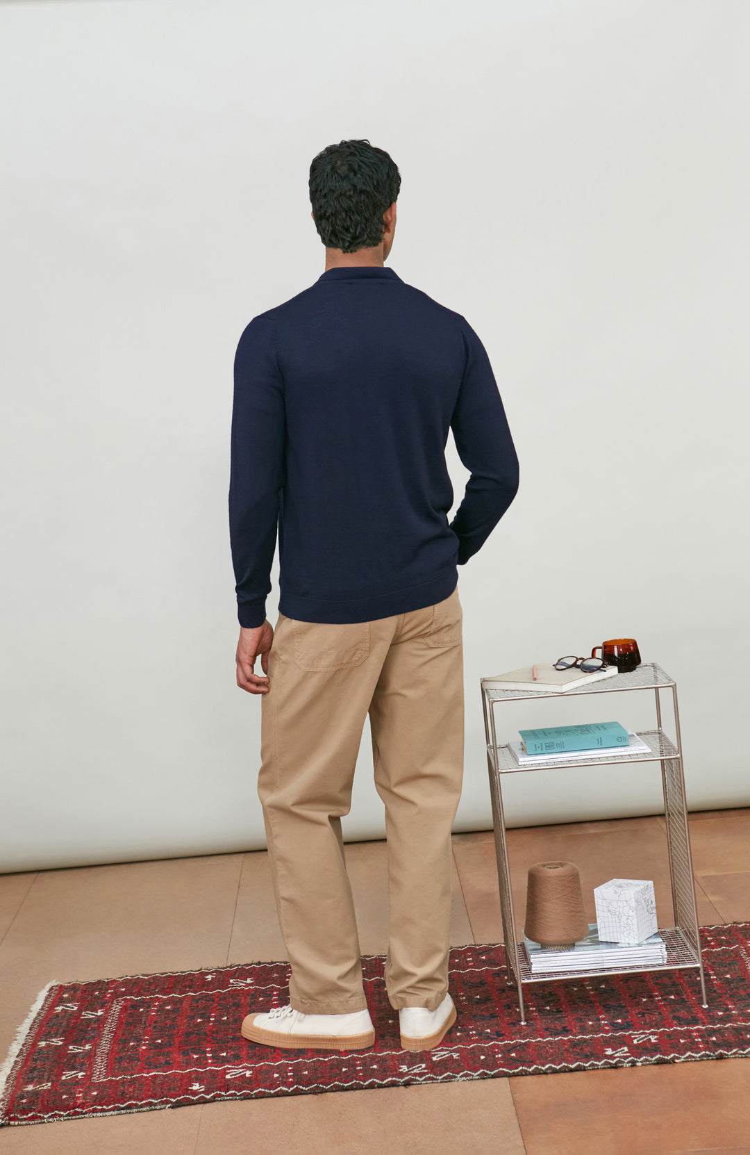 A man wearing a navy blue fine merino wool polo button-up knit cardigan by paul james knitwear over a white undershirt and khaki pants, standing against a plain light gray studio background.