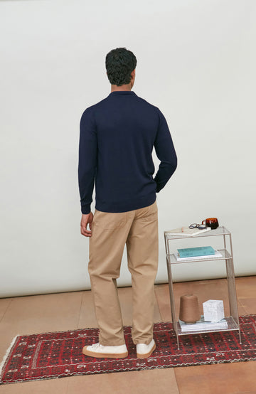A man wearing a navy blue fine merino wool polo button-up knit cardigan by paul james knitwear over a white undershirt and khaki pants, standing against a plain light gray studio background.