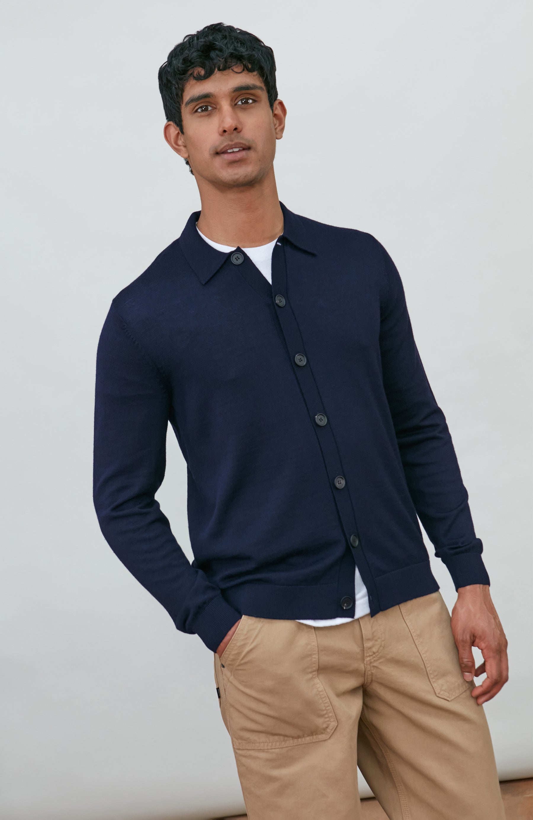 A man wearing a navy blue fine merino wool polo button-up knit cardigan by paul james knitwear over a white undershirt and khaki pants, standing against a plain light gray studio background.