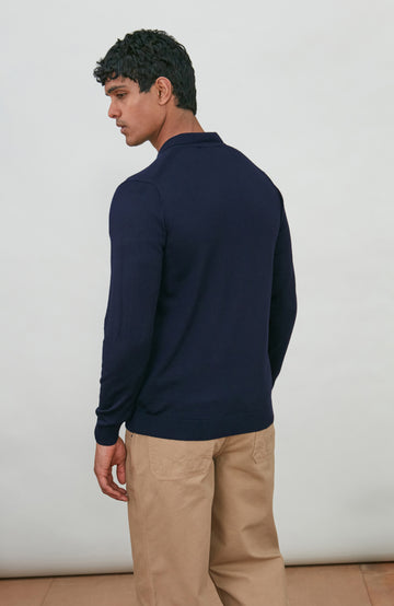 A man wearing a navy blue fine merino wool polo button-up knit cardigan by paul james knitwear over a white undershirt and khaki pants, standing against a plain light gray studio background.