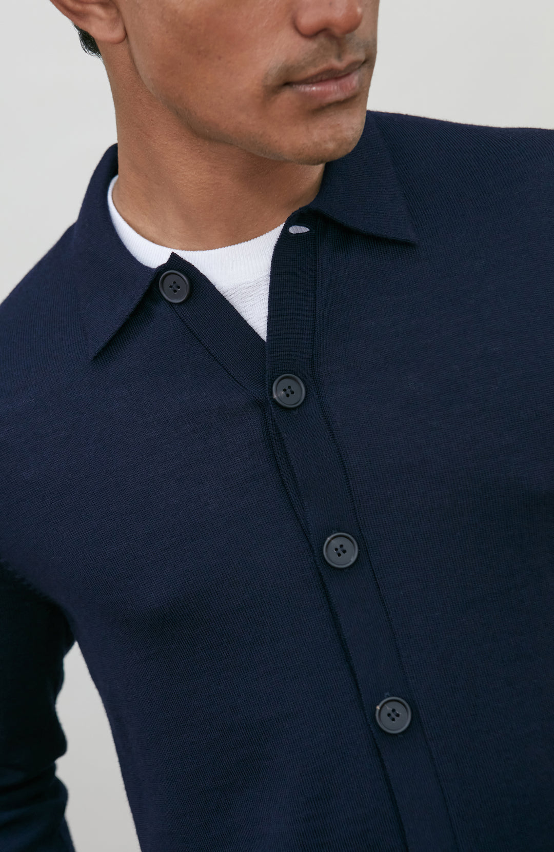 A man wearing a navy blue fine merino wool polo button-up knit cardigan by paul james knitwear over a white undershirt and khaki pants, standing against a plain light gray studio background.
