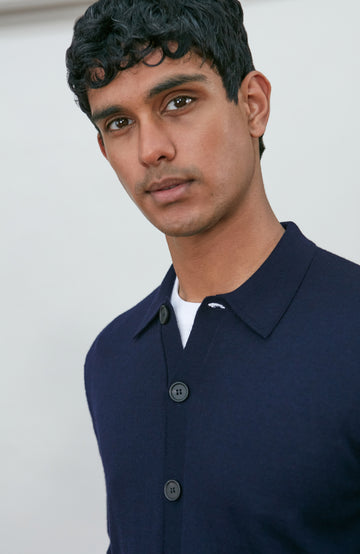 A man wearing a navy blue fine merino wool polo button-up knit cardigan by paul james knitwear over a white undershirt and khaki pants, standing against a plain light gray studio background.