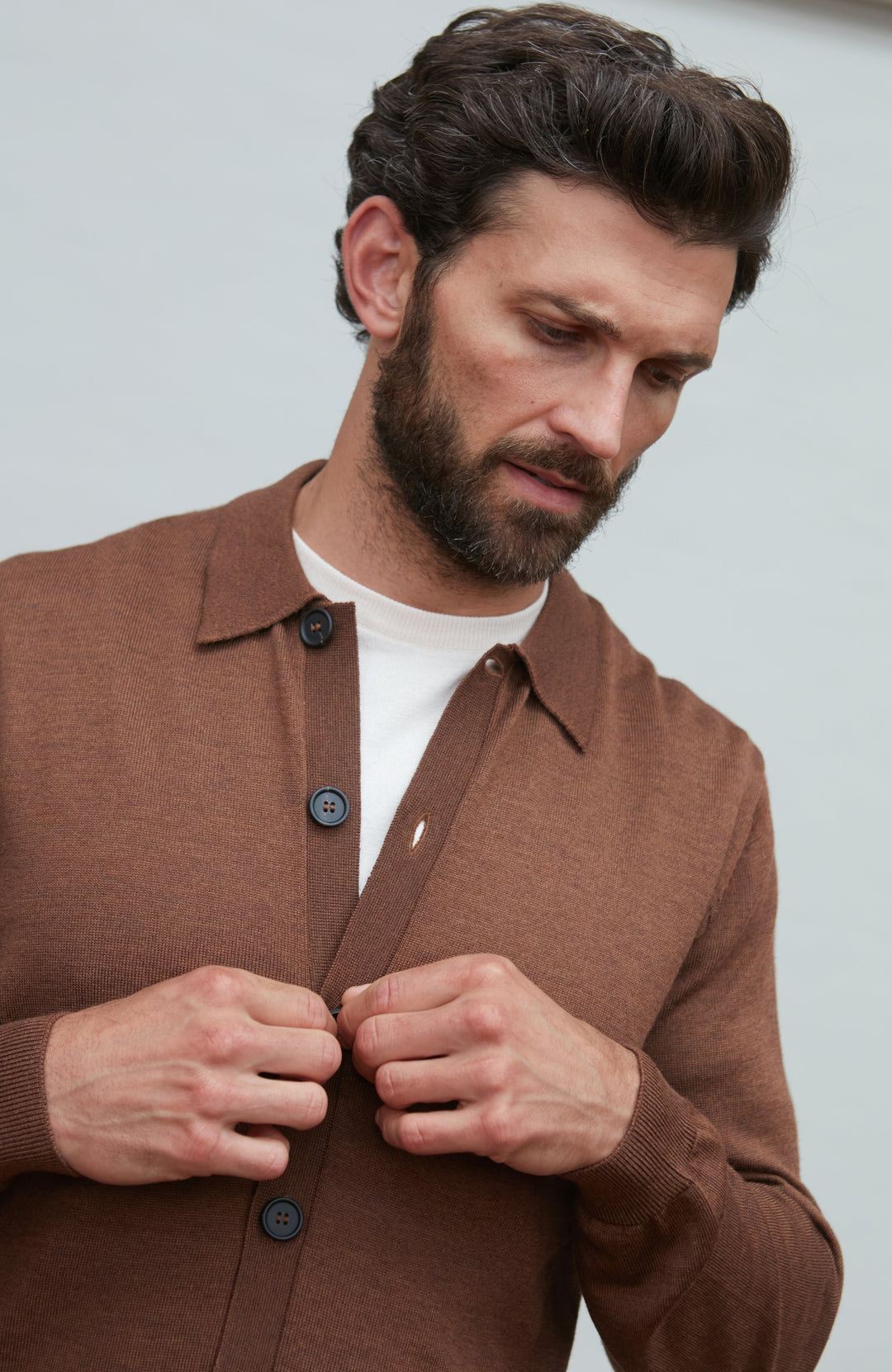 Man wearing a brown fine wool cardigan with a white shirt underneath, against a plain background by Paul James