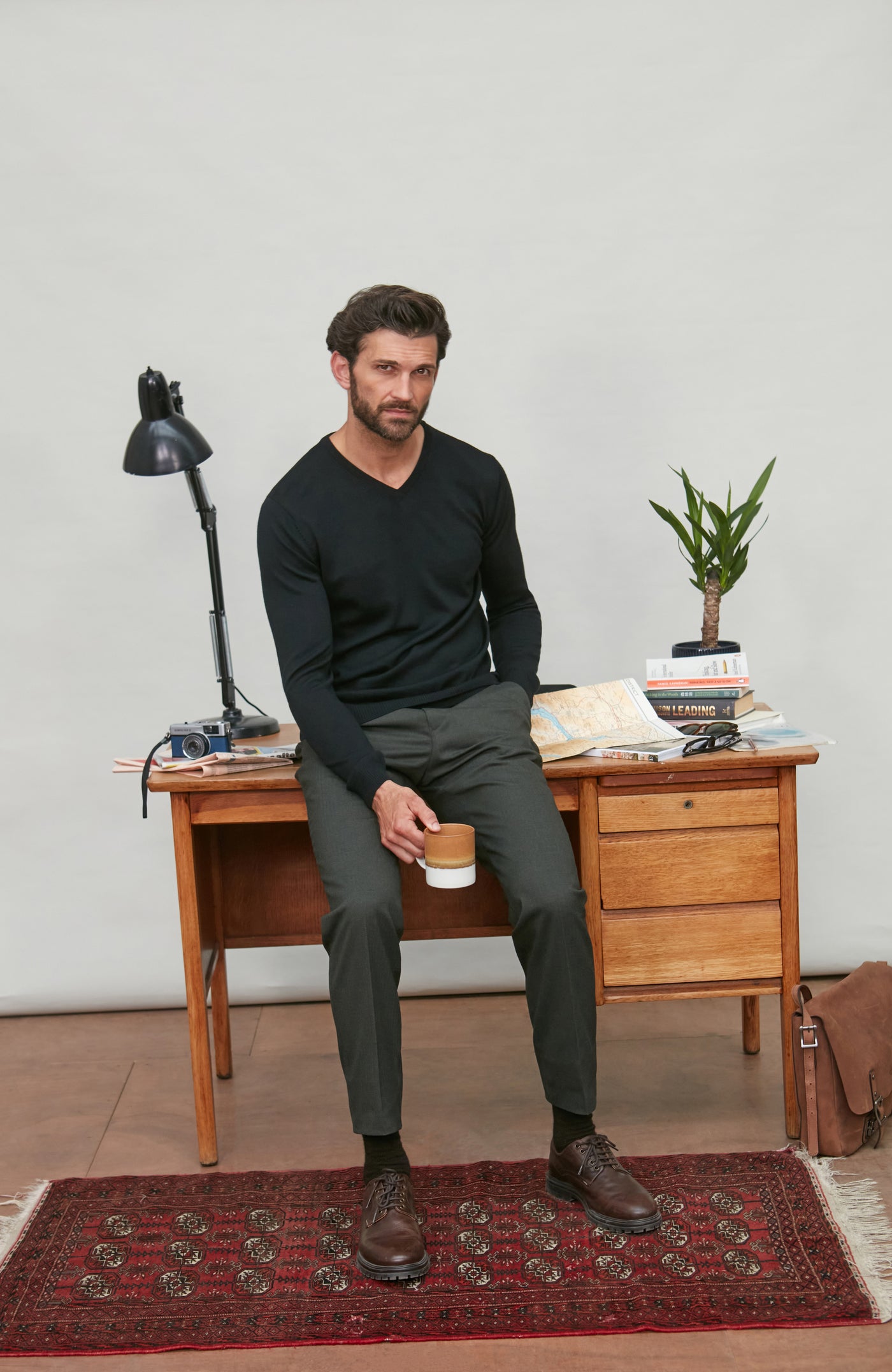 Man in v neck black wool sweater sitting on a wooden desk holding a coffee cup, with a lamp and plant in the background.
