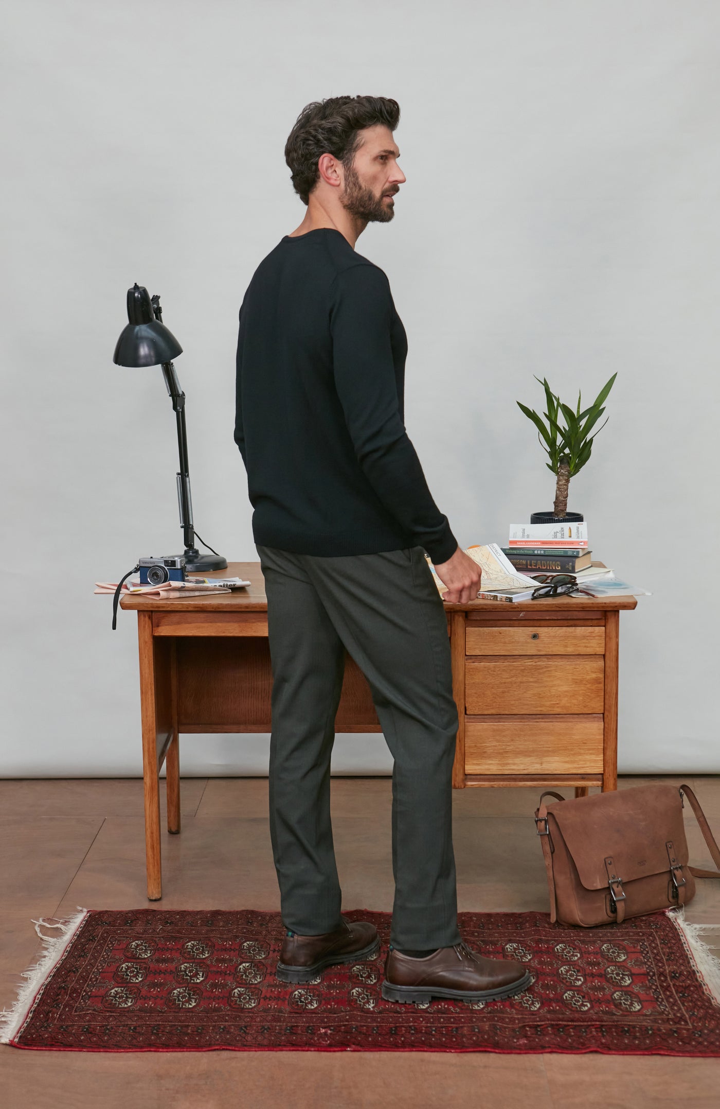 Man wearing black v neck jumper standing in a room with a wooden desk, lamp, and plant.
