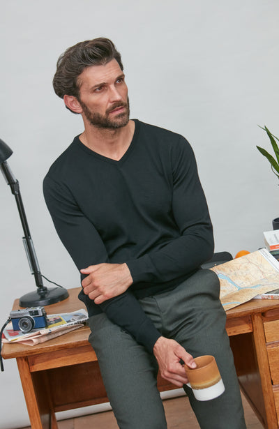 Man wearing a black merino wool v neck sweater and gray pants, sitting at a desk holding a coffee cup, 