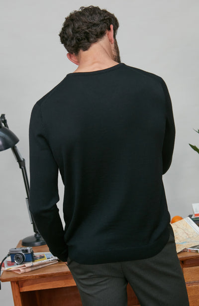 Person wearing a black v merino wool sweater in an indoor setting with a desk and lamp.