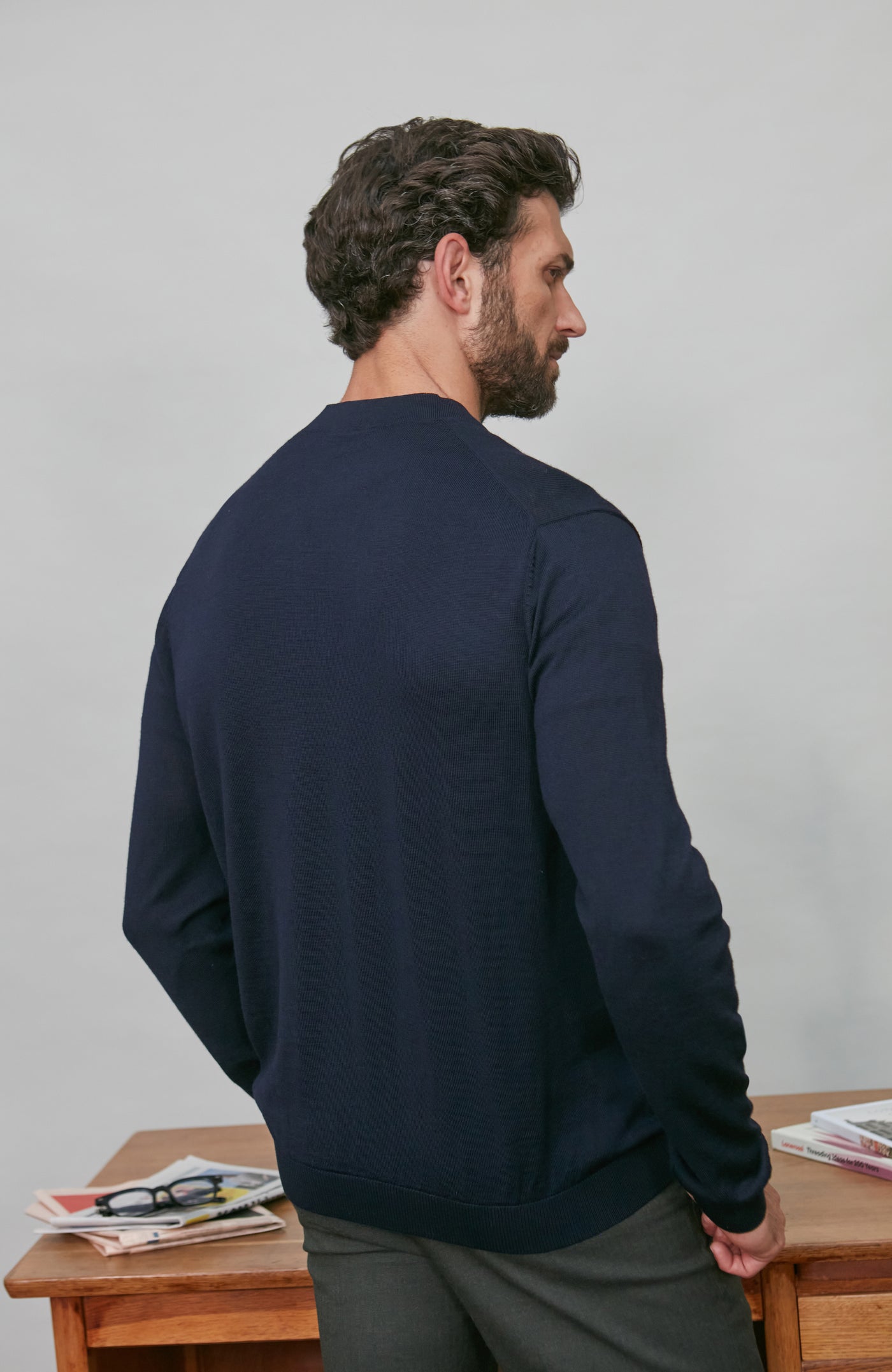 Man wearing a navy blue merino wool cardigan standing in front of a wooden desk with newspapers on it.