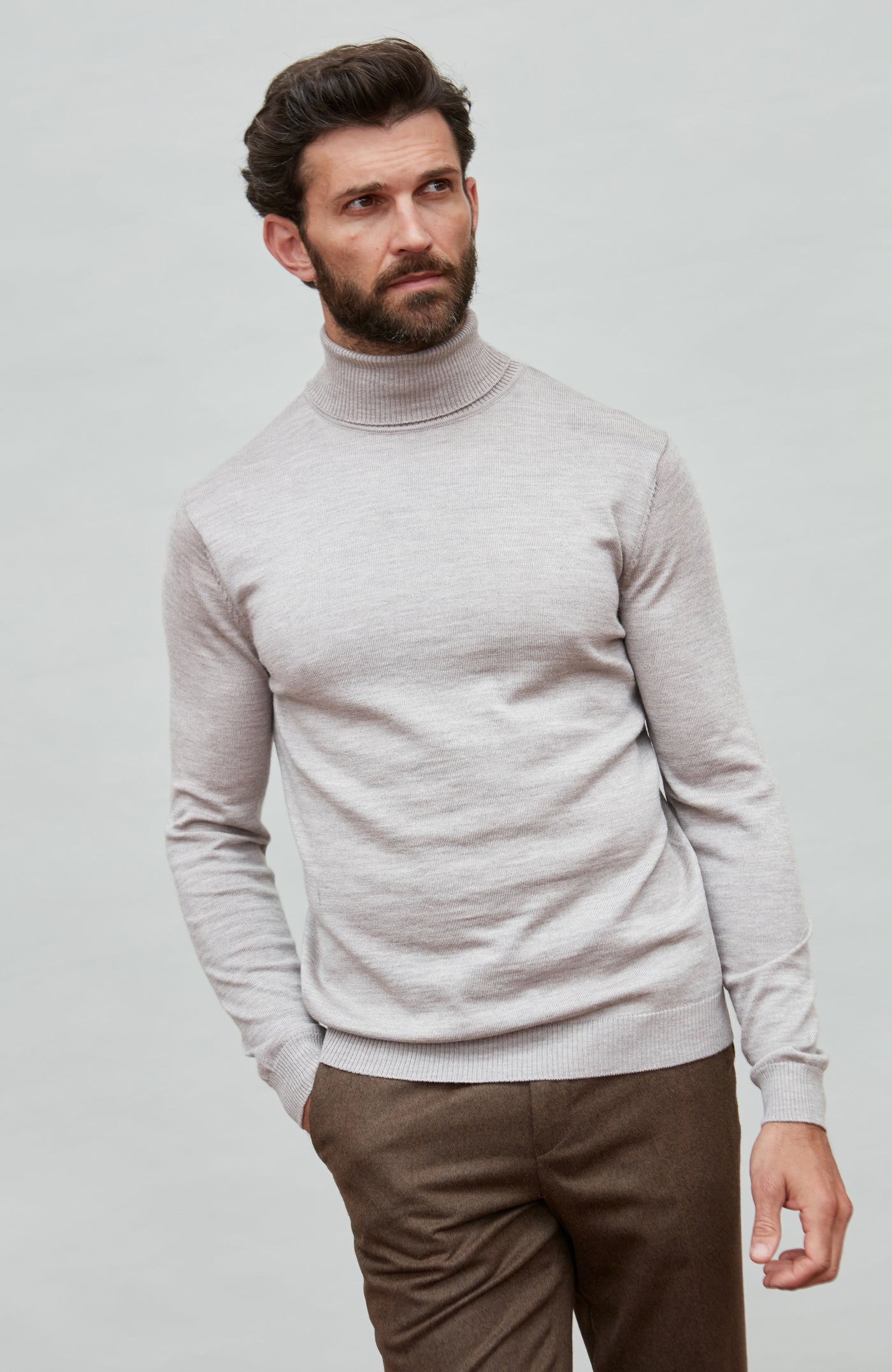 Man wearing a light beige fine merino wool turtleneck sweater and brown pants against a plain background