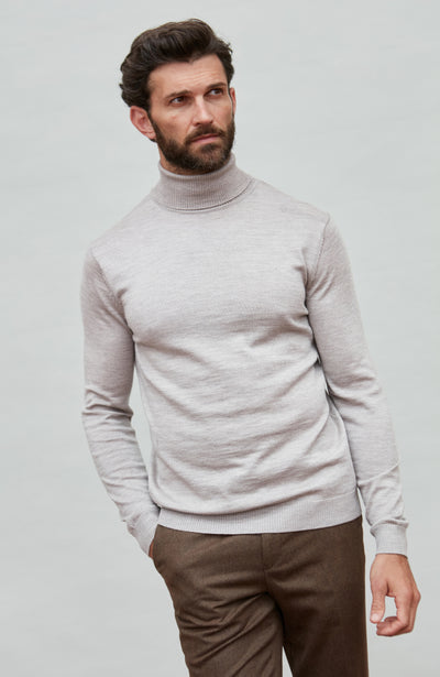 Man wearing a light beige fine merino wool turtleneck sweater and brown pants against a plain background