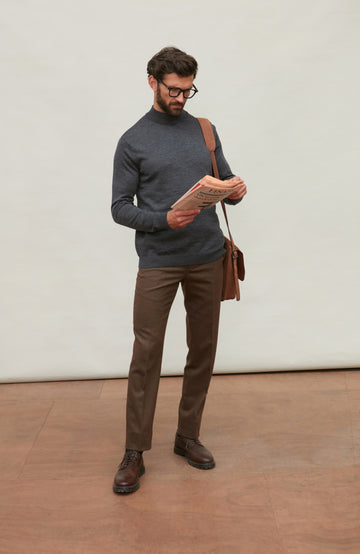 Man wearing a dark gray merino wool turtle neck sweater and brown pants holding a brown leather bag against a light gray background by Paul James
