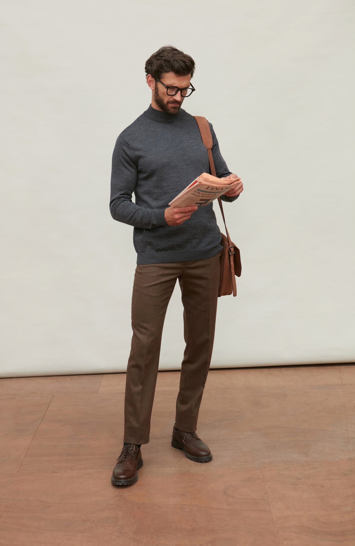 Man wearing a dark gray merino wool turtle neck sweater and brown pants holding a brown leather bag against a light gray background