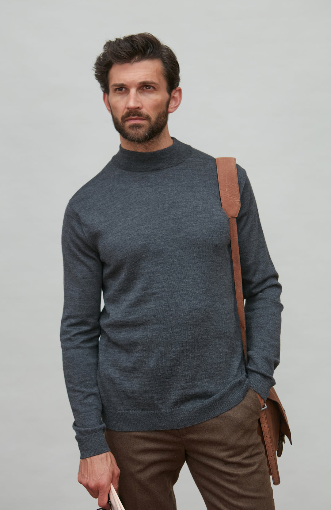 Man wearing a dark gray merino wool turtle neck sweater and brown pants holding a brown leather bag against a light gray background by Paul James