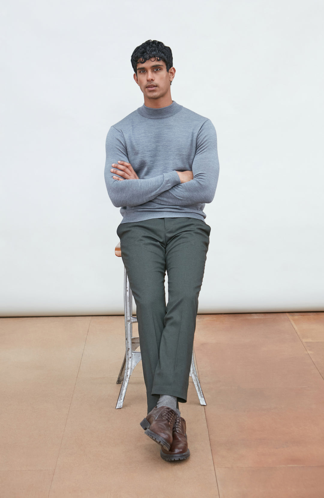 A man wearing a light gray fine merino wool ribbed mock neck sweater by paul james knitwear and dark gray trousers, leaning on a wooden chair against a plain off-white background.