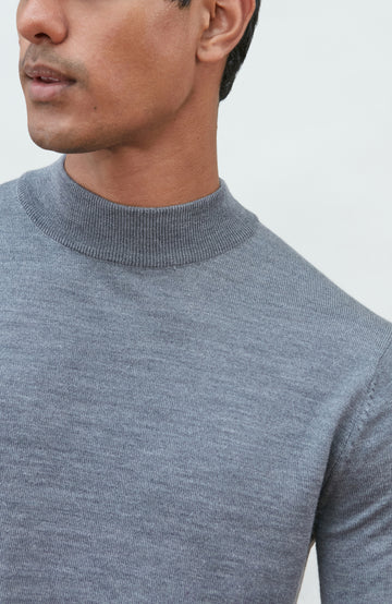 A man wearing a light gray fine merino wool ribbed mock neck sweater by paul james knitwear and dark gray trousers, leaning on a wooden chair against a plain off-white background.