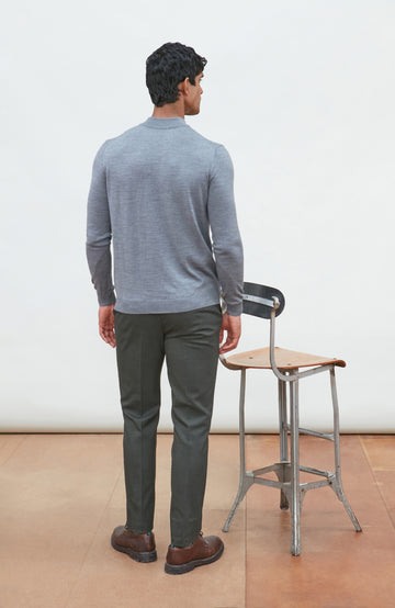 A man wearing a light gray fine merino wool ribbed mock neck sweater by paul james knitwear and dark gray trousers, leaning on a wooden chair against a plain off-white background.