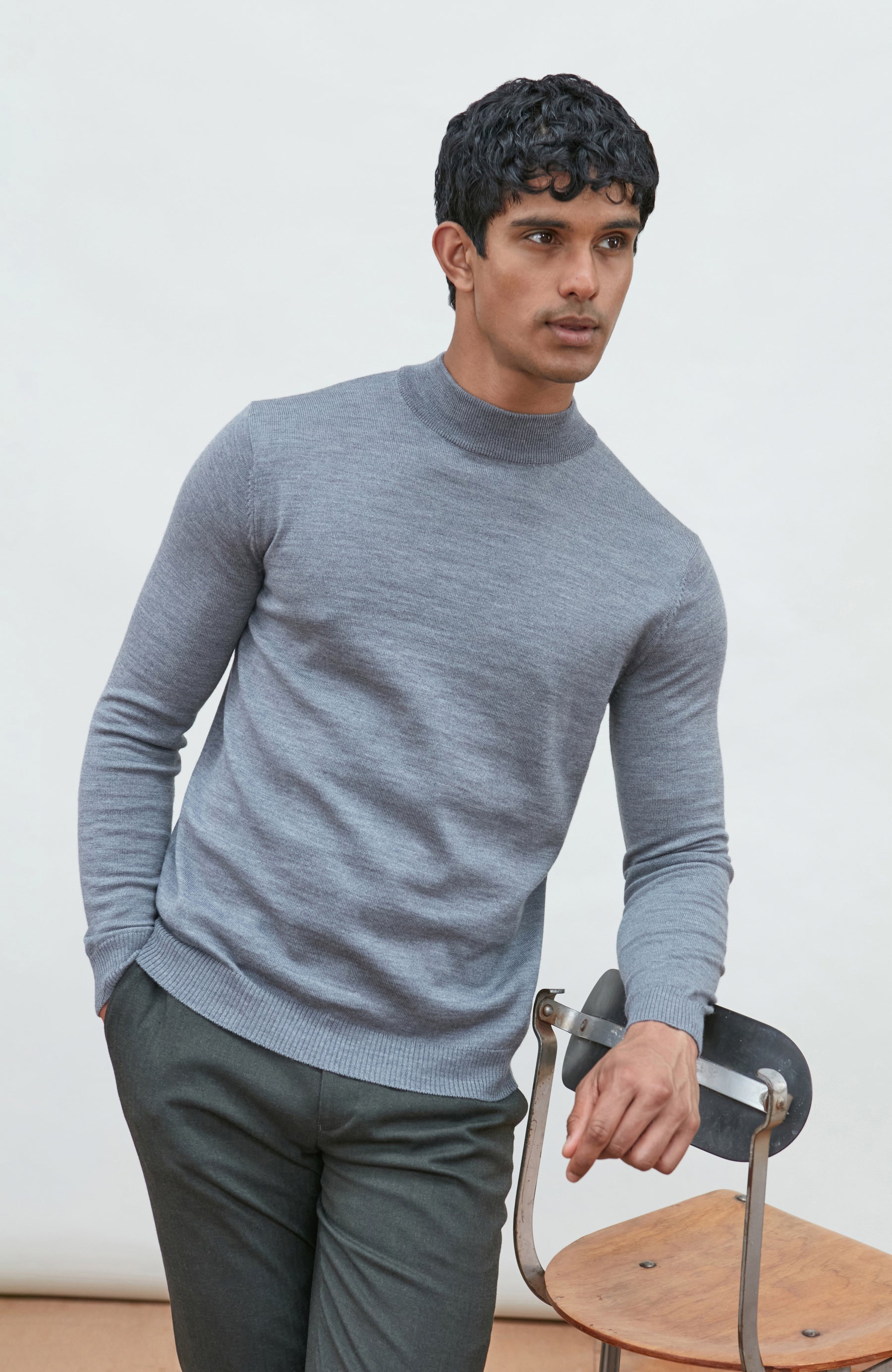 A man wearing a light gray fine merino wool ribbed mock neck sweater by paul james knitwear and dark gray trousers, leaning on a wooden chair against a plain off-white background.