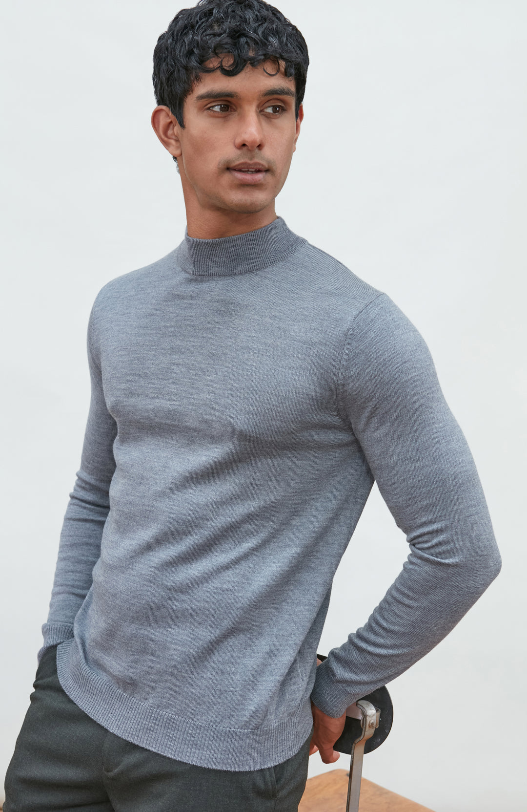 A man wearing a light gray fine merino wool ribbed mock neck sweater by paul james knitwear and dark gray trousers, leaning on a wooden chair against a plain off-white background.