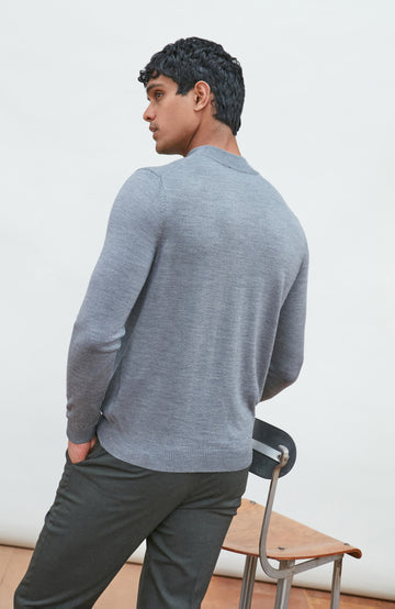A man wearing a light gray fine merino wool ribbed mock neck sweater by paul james knitwear and dark gray trousers, leaning on a wooden chair against a plain off-white background.